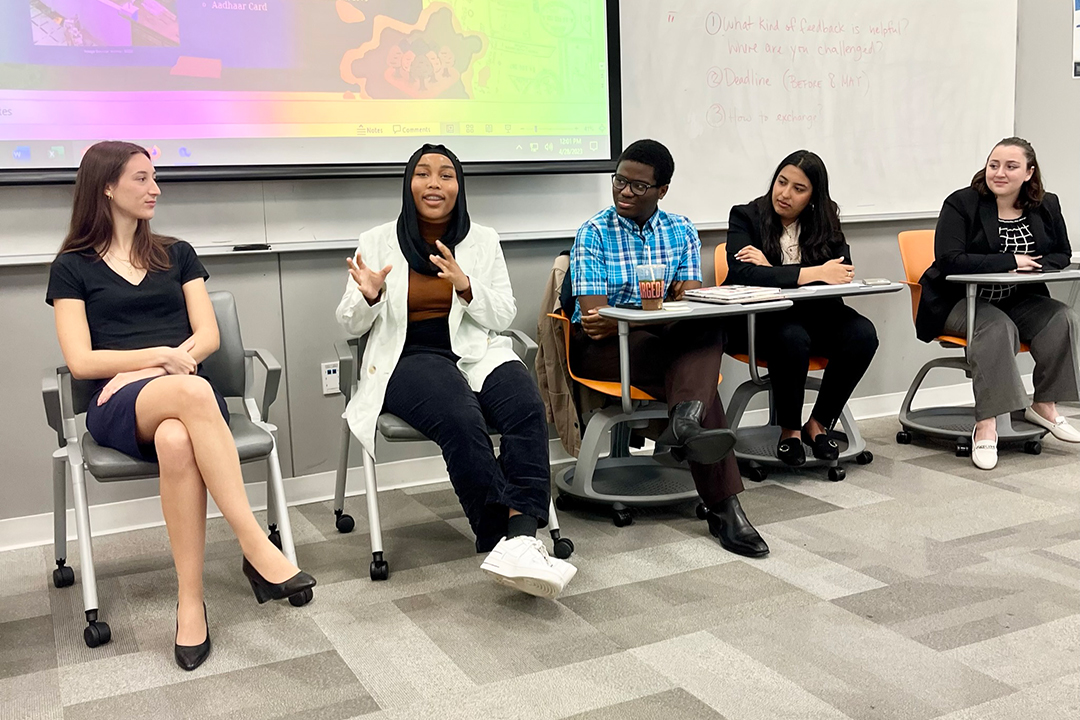 Five students seated at desks at the front of a classroom gesturing and presenting to the room