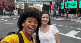 Two first-year GW students taking a selfie in DC's Chinatown, with neon signs, a painted Chinese gate, and crosswalks visible in the background